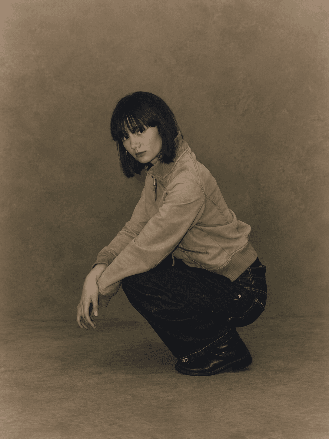 A woman crouching in a tan leather jacket and dark R/K2 denim against a warm studio backdrop