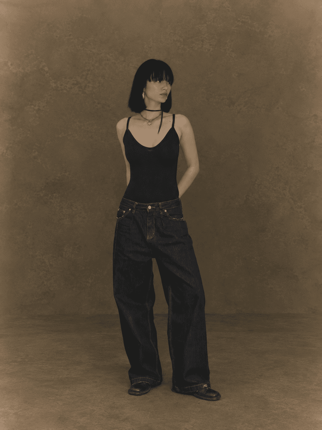 A woman in a black top and wide dark R/K2 denim against a warm studio backdrop