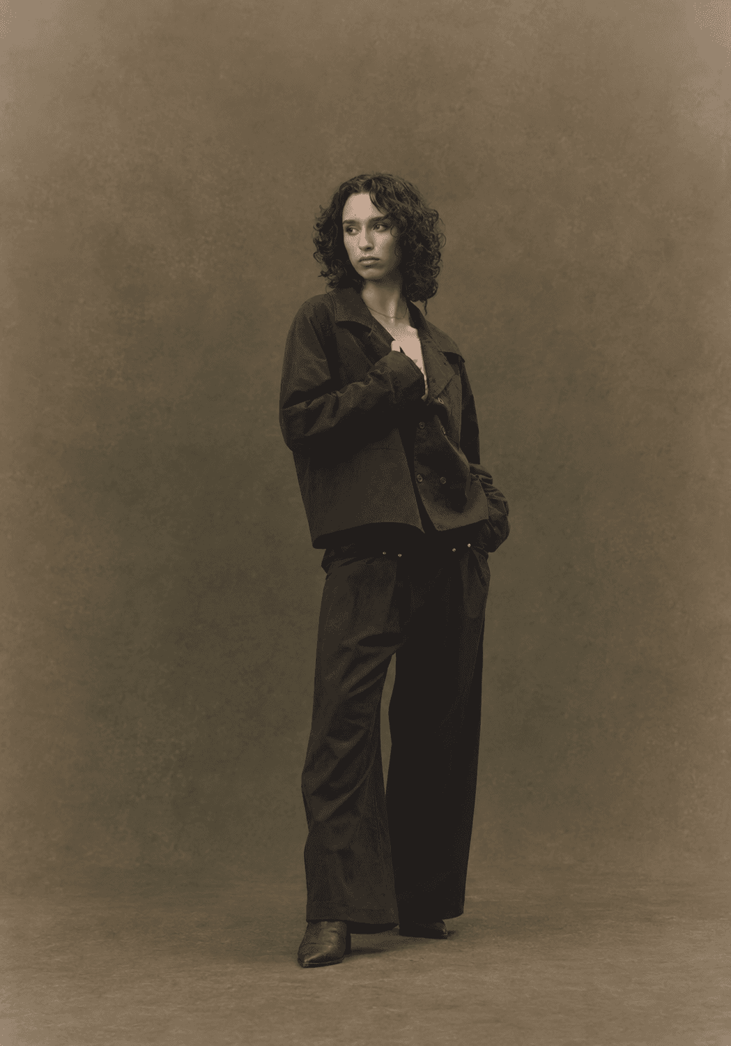A woman in a dark R/K2 jacket and wide trousers against a warm studio backdrop