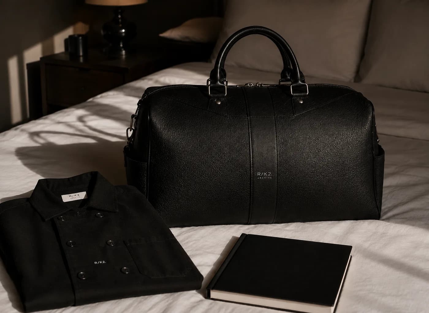 A black R/K2 leather weekend bag on a bed beside a jacket and a book